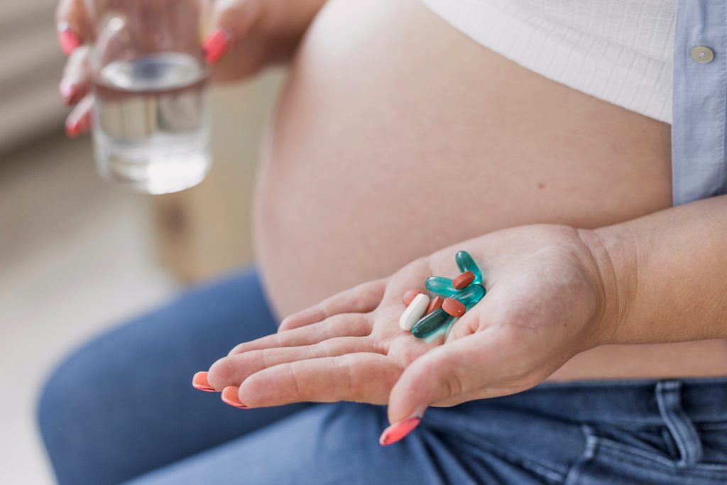 Pregnant woman holding a glass of water and pills, looking thoughtfully at the medication in her hand